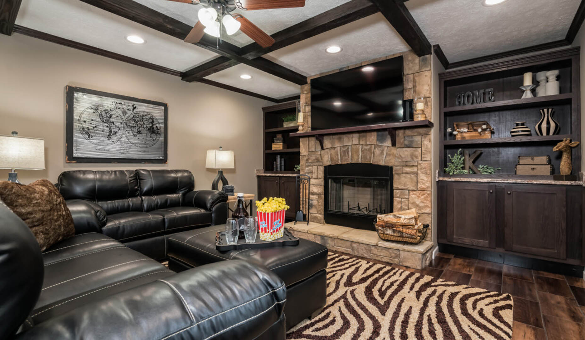 Cozy living room with black leather sofas, a stone fireplace, and zebra-patterned rug. A ceiling fan and warm lighting create an inviting atmosphere.