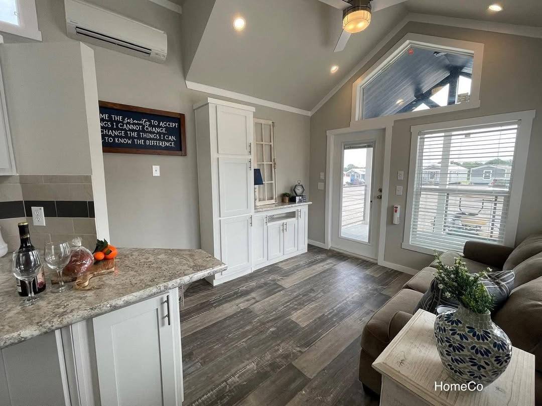 Modern cozy living room with vaulted ceiling and bright natural light. Features white cabinets, a gray marble countertop, and wood flooring. A sofa and decorative vase add comfort and elegance.