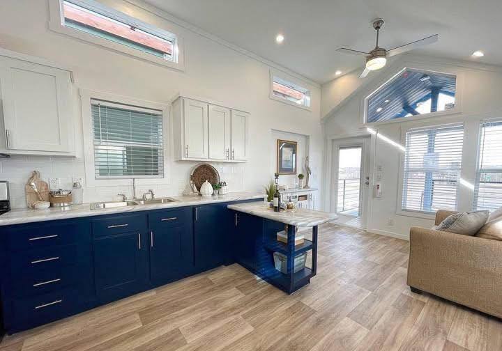 Bright, modern kitchen with blue and white cabinets, wood flooring, and a ceiling fan. Large windows enhance the airy atmosphere, with a cozy beige couch nearby.