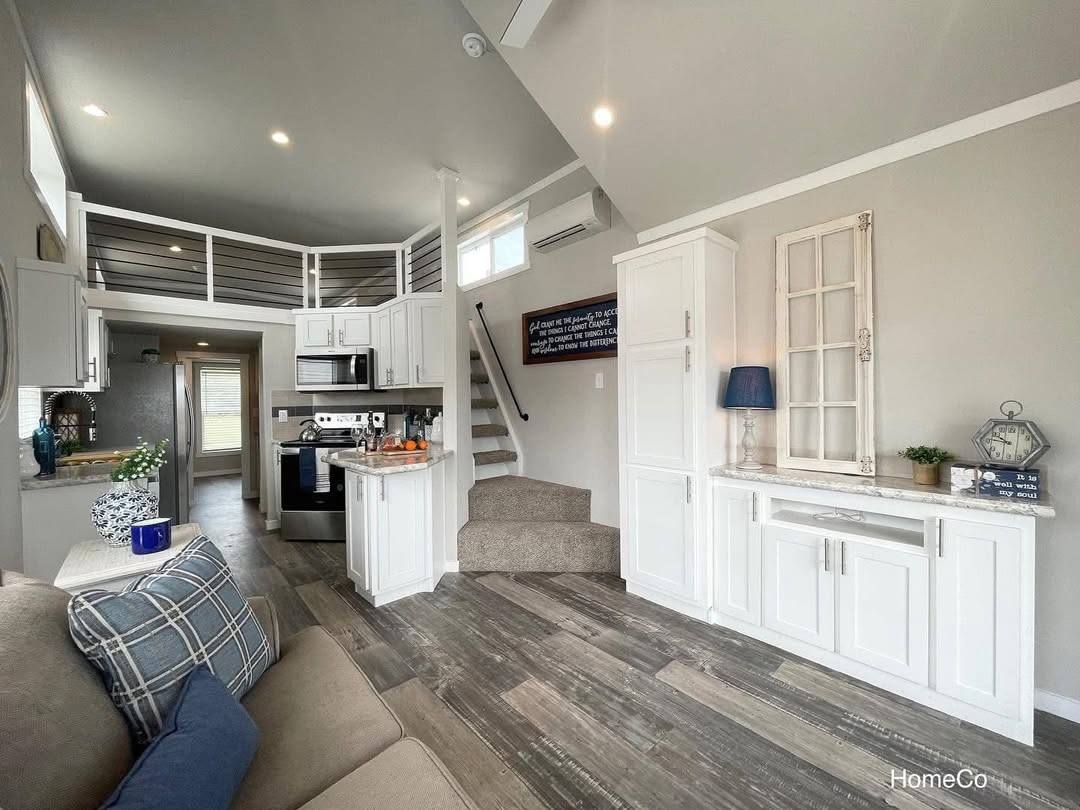 Modern tiny home interior with wooden floors, white cabinets, and vaulted ceiling. Cozy loft, compact kitchen, stairs, and calming neutral tones.