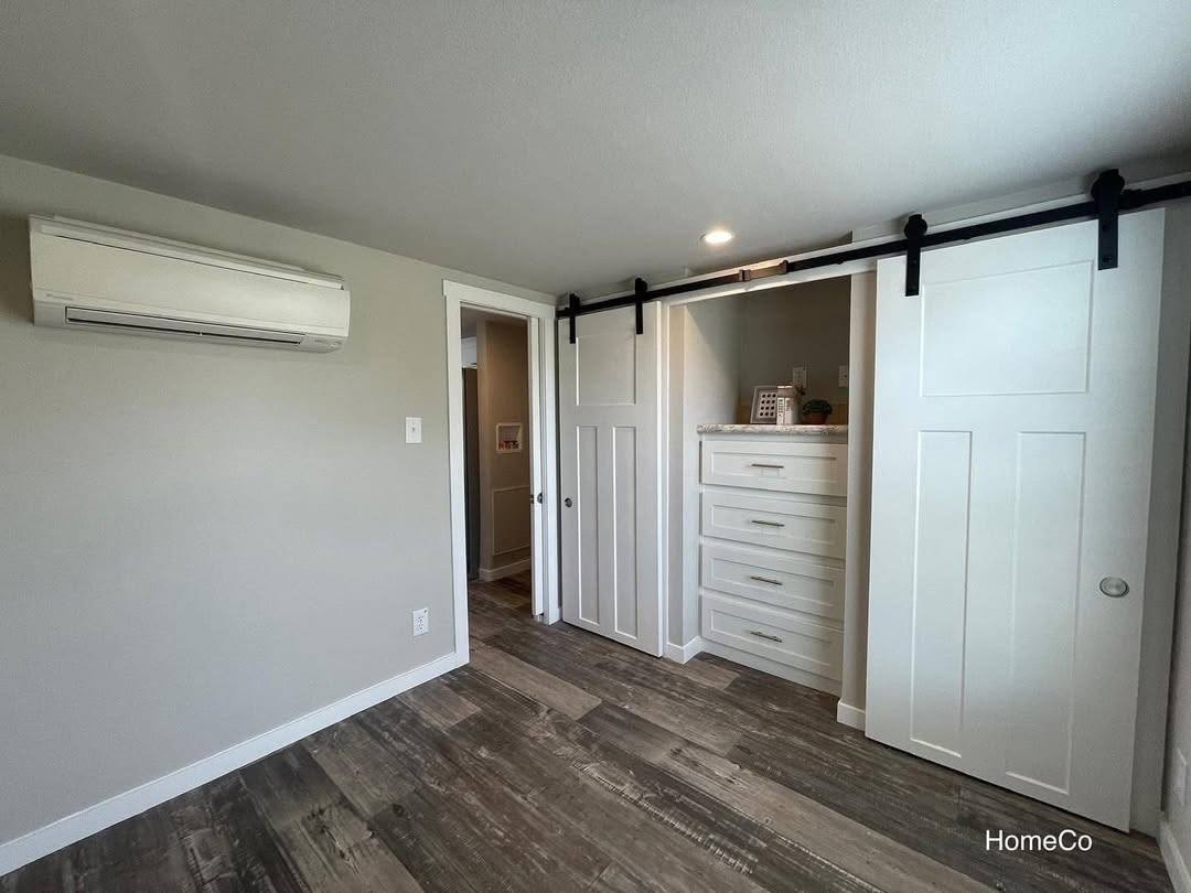 Minimalist room with light gray walls, featuring a white air conditioning unit and a closet with white sliding barn doors. Dark wood flooring adds contrast.