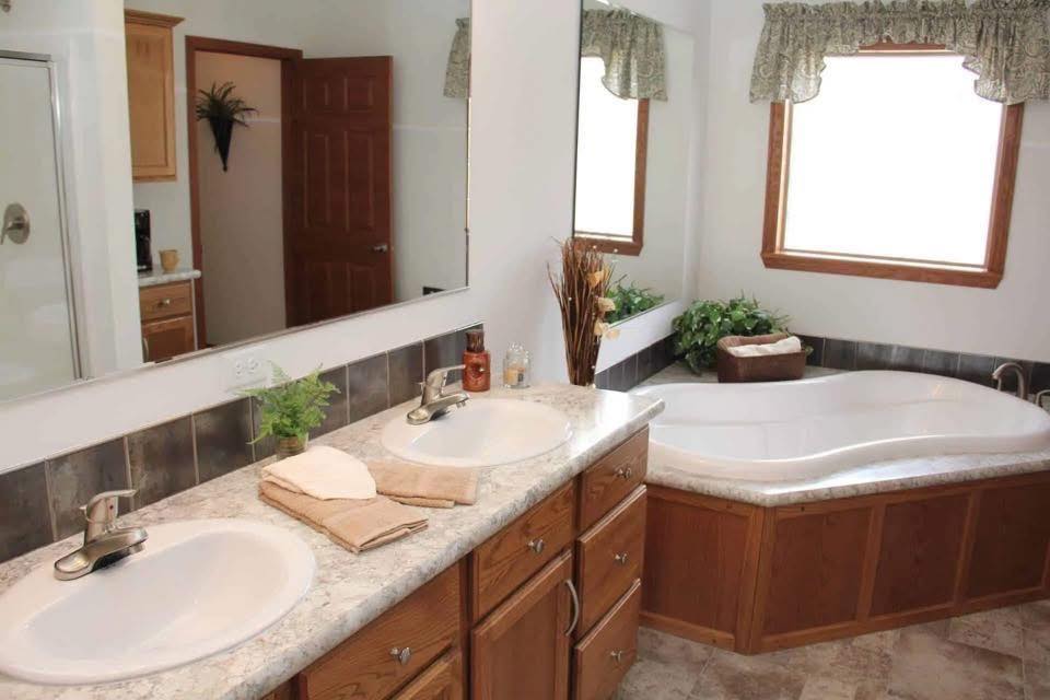 Bright bathroom with a curved bathtub, large window with green curtains, dual sinks, and a mirror. Decor includes plants and towels, conveying a cozy, serene vibe.