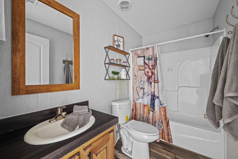 Cozy bathroom with a white sink and wooden vanity, a mirror, and a decorative shower curtain. Shelves hold plants, conveying a warm, rustic feel.