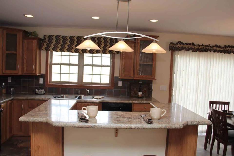 A cozy kitchen with brown cabinets and granite countertops. A central island holds two mugs and napkins. Ambient light from a window and lamp adds warmth.