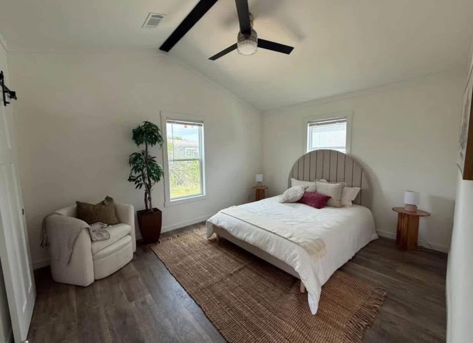 Minimalist bedroom with a large bed, decorative cushions, and a round headboard. A potted plant and armchair add a cozy touch. Warm tones and soft lighting create a serene ambiance.