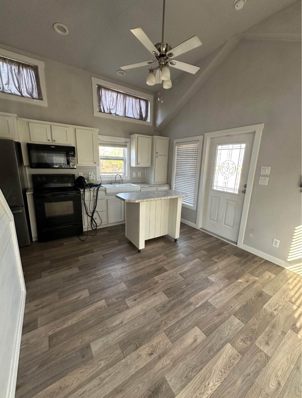 Compact kitchen with white cabinets, dark appliances, and light countertops. A small island sits on wood flooring. Ceiling fan overhead, bright natural light.