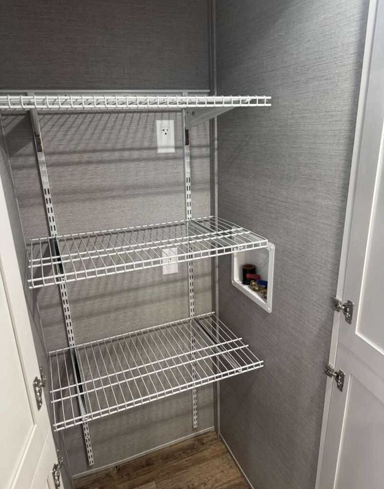 Empty pantry with three wire shelves on a textured gray wall, and a small inset alcove on the right, containing a few small bottles.