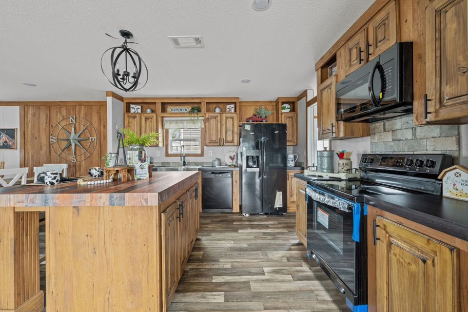 Rustic kitchen with wooden cabinets and a large island. Features a black fridge, stove, and microwave. Earthy tones and farmhouse décor create a cozy feel.