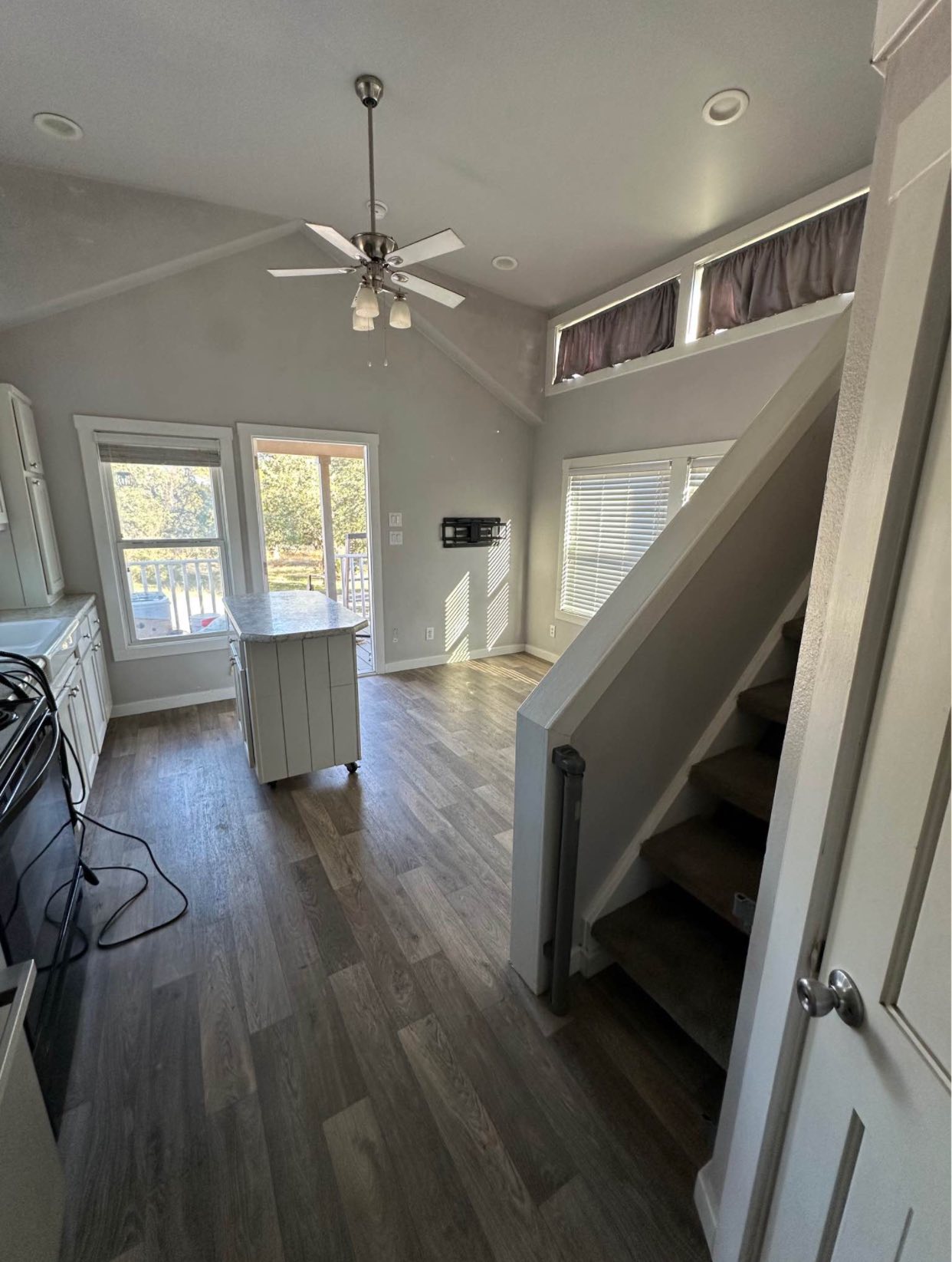 Modern interior with high ceiling, ceiling fan, and natural light. A staircase leads upstairs. Wooden floor, kitchen with island, white cabinets. Cozy and bright.