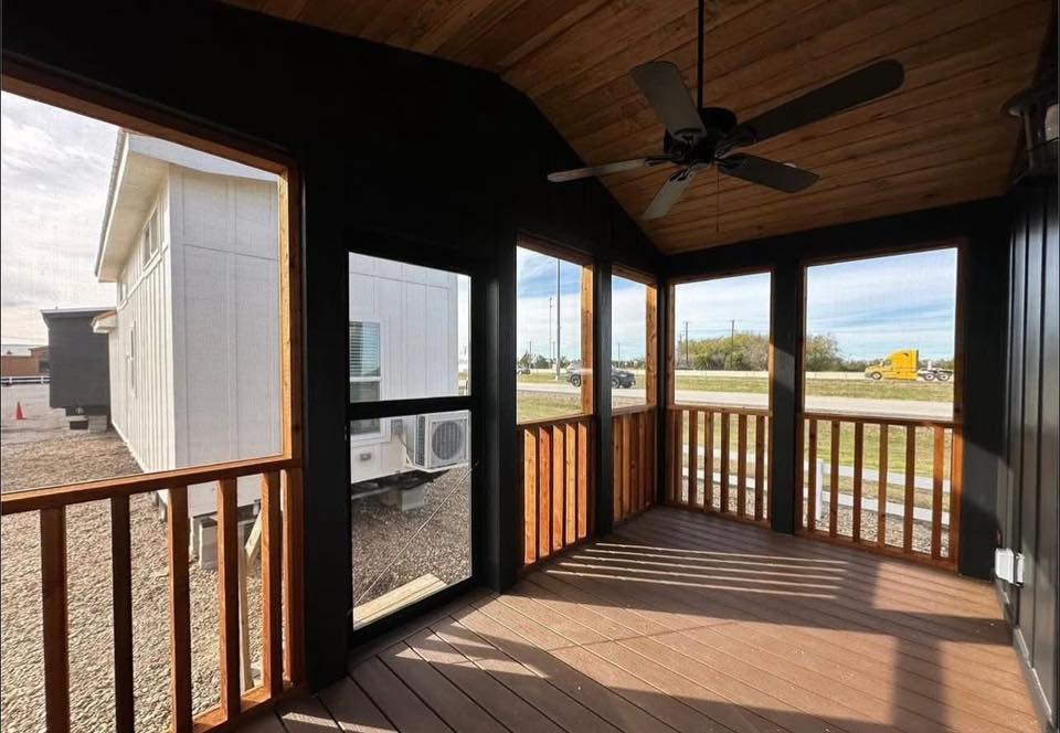 A screened porch with wooden floors and ceiling fan, surrounded by wooden railing. View of a neighboring house and distant road with a truck. Bright and airy.