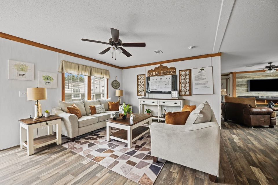 Cozy farmhouse-style living room with beige sofas, brown cushions, a wooden coffee table, and rustic decor. A ceiling fan and natural light add warmth.
