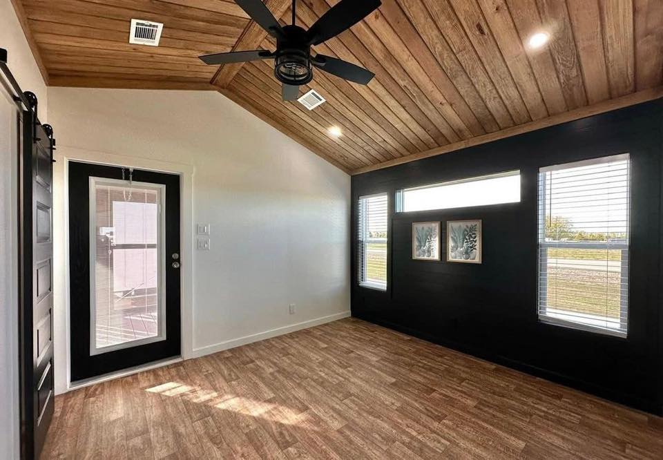 Modern room with wooden ceiling and floor, black accent wall, and three windows. A black ceiling fan hangs above. Artwork adds a touch of elegance.