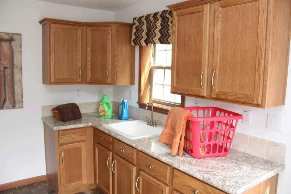 A laundry room with wooden cabinets, a marble countertop, sink, detergent bottles, a red laundry basket, and orange towels. Sunlight filters through a window.