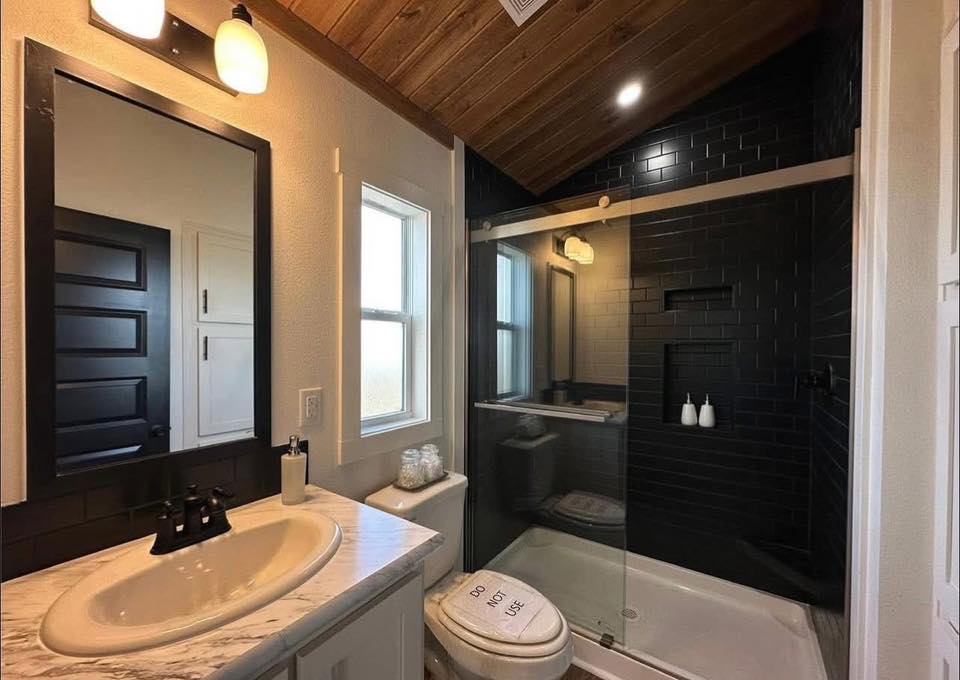 Cozy bathroom with wood ceiling, black tiled shower, and glass door. White countertop with sink, mirror, and note on toilet seat reading “Do Not Use.”