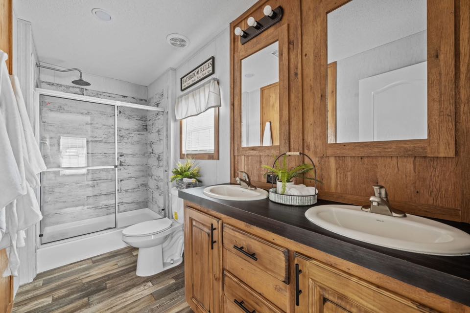 A rustic bathroom with wooden cabinetry, dual sinks, and mirrors. A large shower with sliding doors is opposite a toilet, under a sign and window.