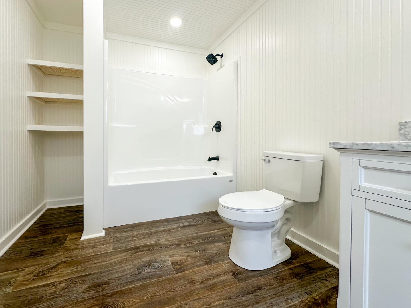 A bright, modern bathroom featuring a white bathtub with black fixtures, white toilet, wood floor, and wood shelving. The overall tone is clean and minimalist.