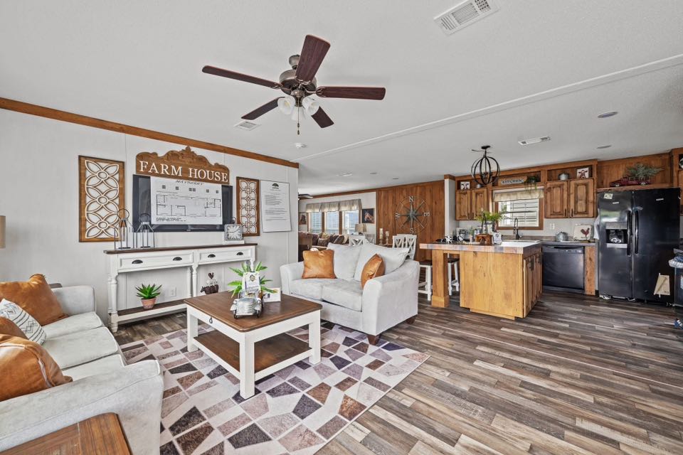 Warm farmhouse-style living room and kitchen. Features cozy sofas, wooden accents, a ceiling fan, modern decor, and a rustic ambiance.