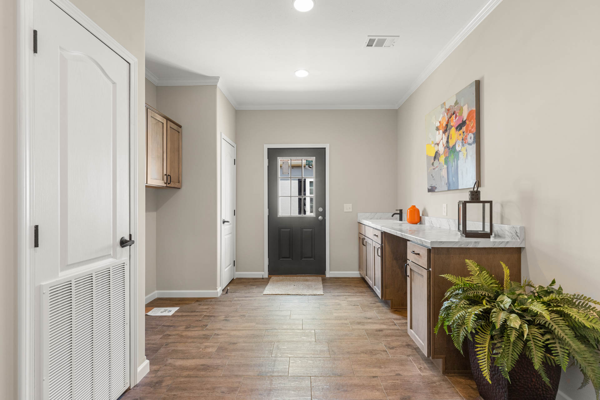 A bright mudroom with wood flooring, white door, and a dark entrance. Features a counter with decorative items and a plant; colorful wall art above.
