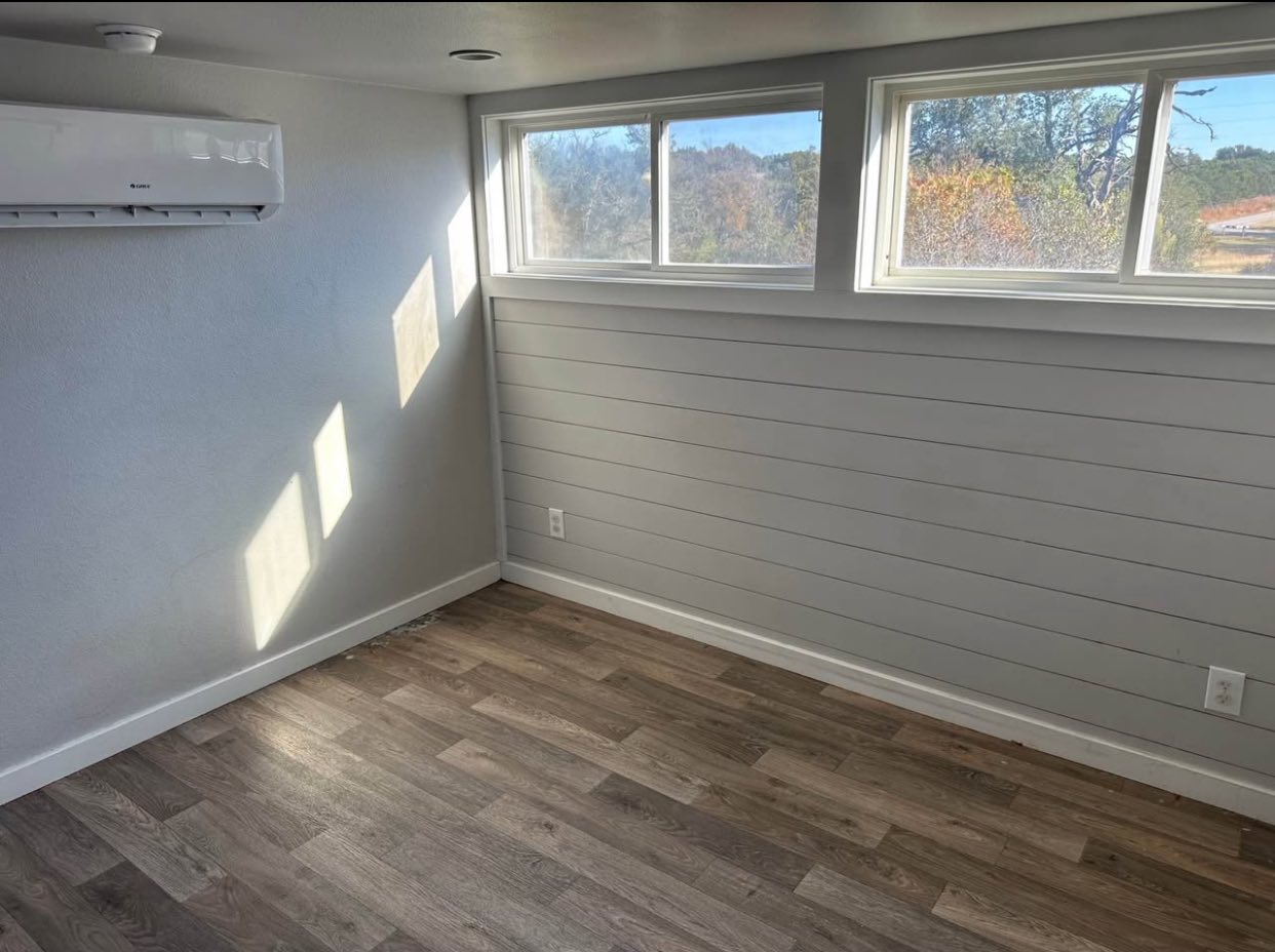Small, sunlit room with light wood flooring, white shiplap walls, narrow horizontal windows, and a mounted air conditioning unit. Bright and minimal.