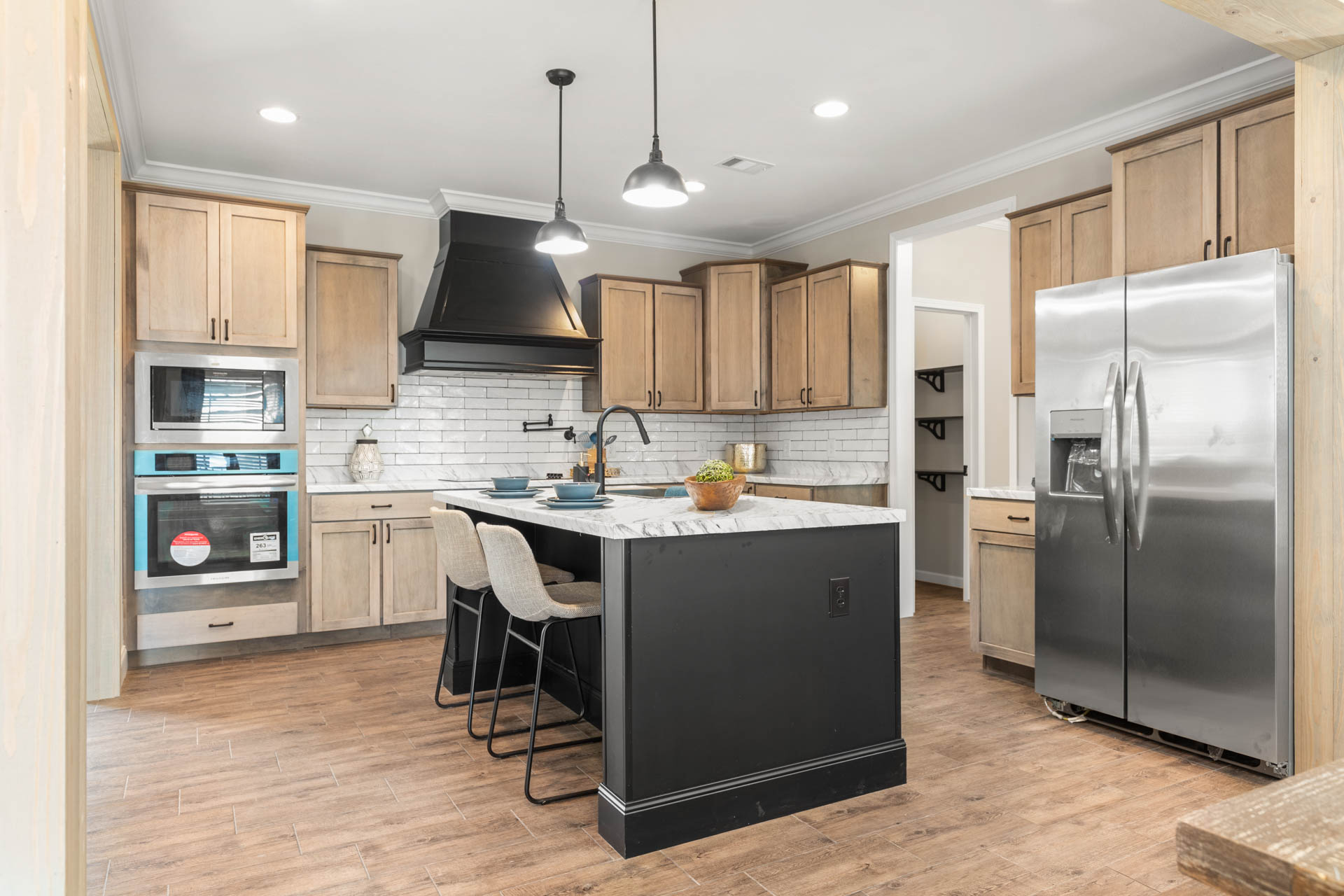 Modern kitchen with light wood cabinets, black range hood, and stainless steel fridge. Features marble island with stools, pendant lighting, and wood floors.
