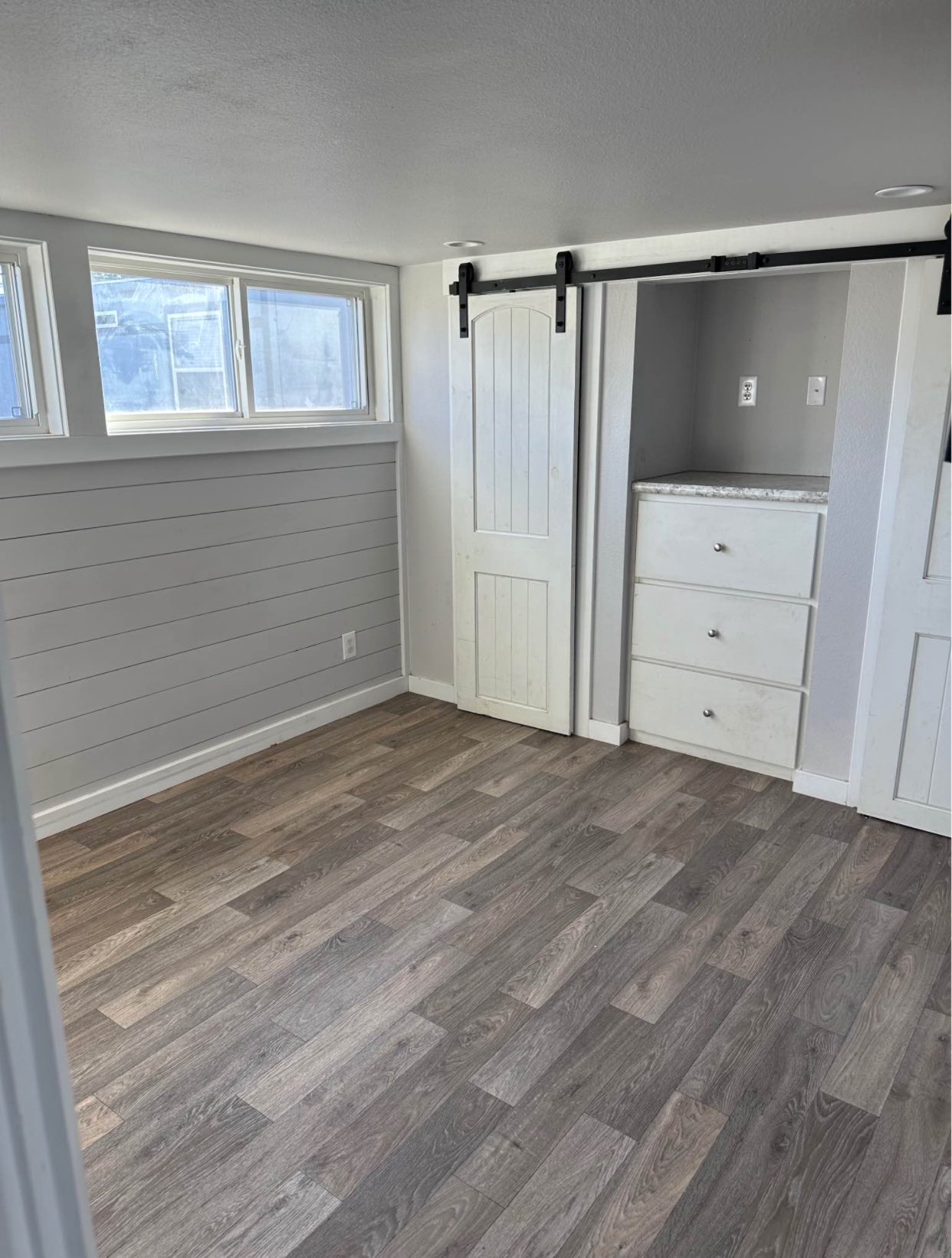 Small room with gray wood flooring, light gray walls, and a bright window. Features a sliding barn door beside white drawers, creating a cozy, modern feel.