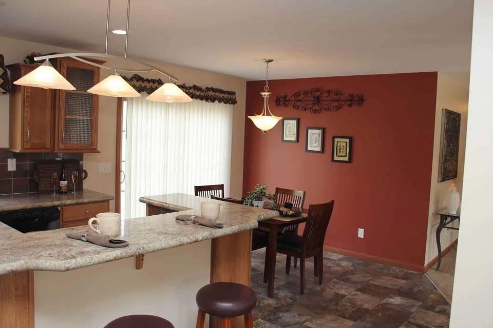 A kitchen with a granite breakfast bar and two stools is lit by pendant lights. In the background, a dining area with a red accent wall and framed art creates a cozy, inviting atmosphere.