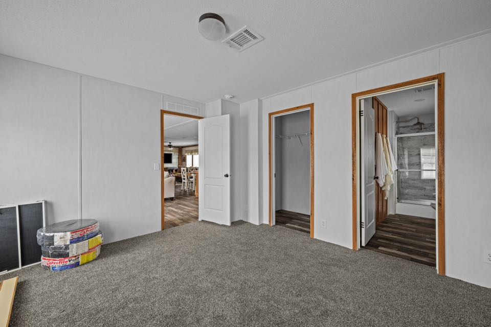 Empty room with gray carpet, white walls, and wooden door frames. Two open doorways lead to a bathroom and a closet. Rolls of insulation rest in the corner.