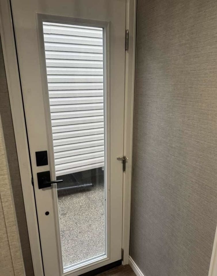 A white door with a narrow vertical window, revealing an outdoor view of siding and gravel. The indoor wall is textured, conveying a modern tone.