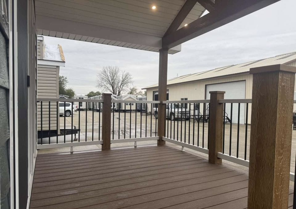 Covered porch with wood flooring and railings overlooking a rainy industrial area with parked vehicles, warehouses, and a distant tree.