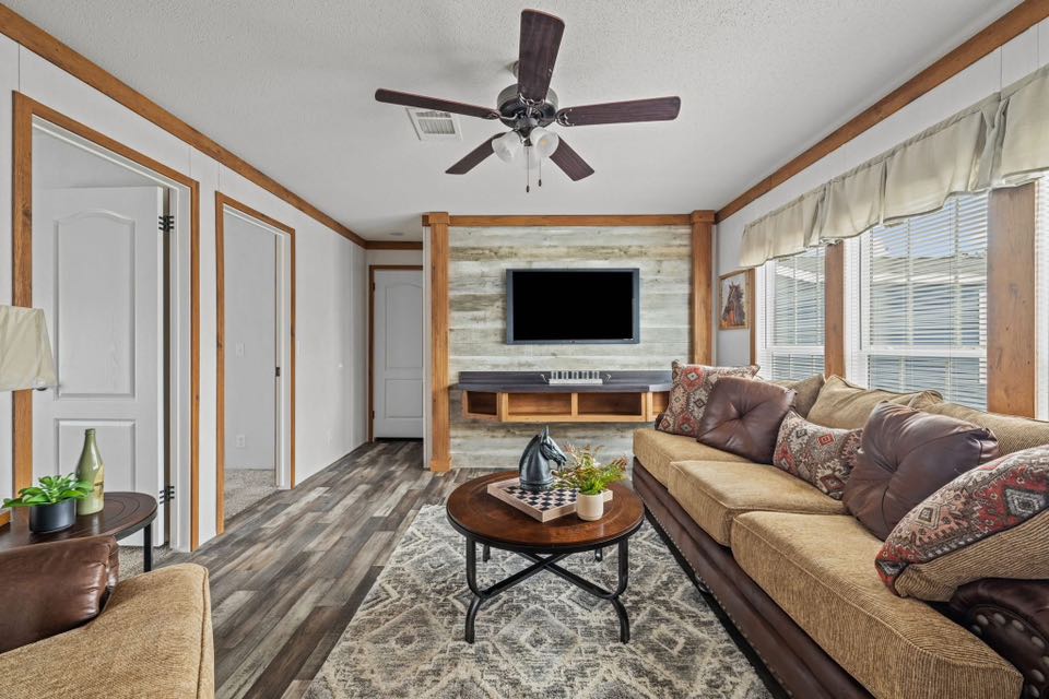 Cozy living room with beige sofas, patterned cushions, a wooden coffee table holding a plant and books, a wall-mounted TV, ceiling fan, and large windows.