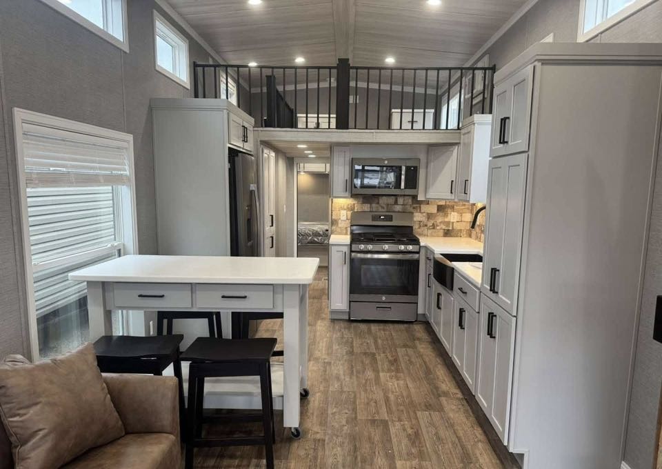 Modern tiny house interior with open layout. Features a kitchen with white cabinets, stainless steel appliances, loft railing above, and cozy couch.