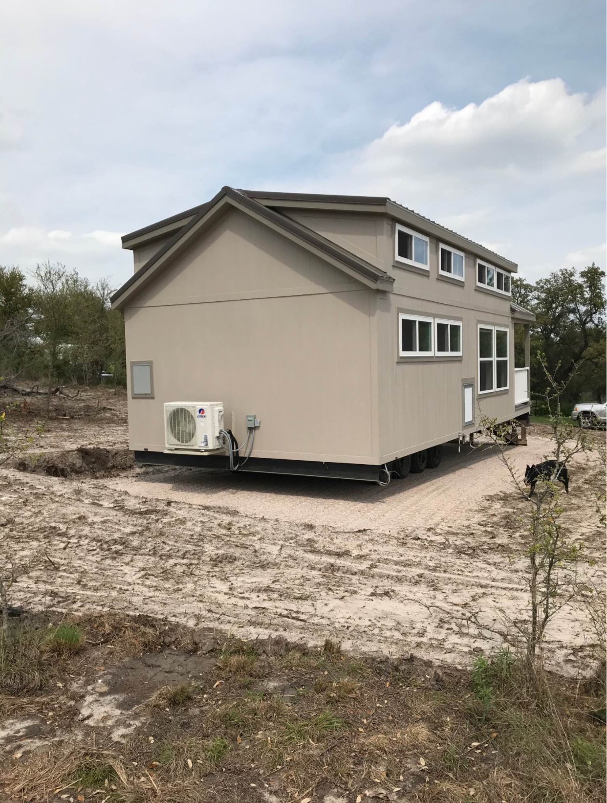 A beige tiny house sits on wheels on a cleared plot of land under a cloudy sky. It features multiple windows and a small air conditioning unit.