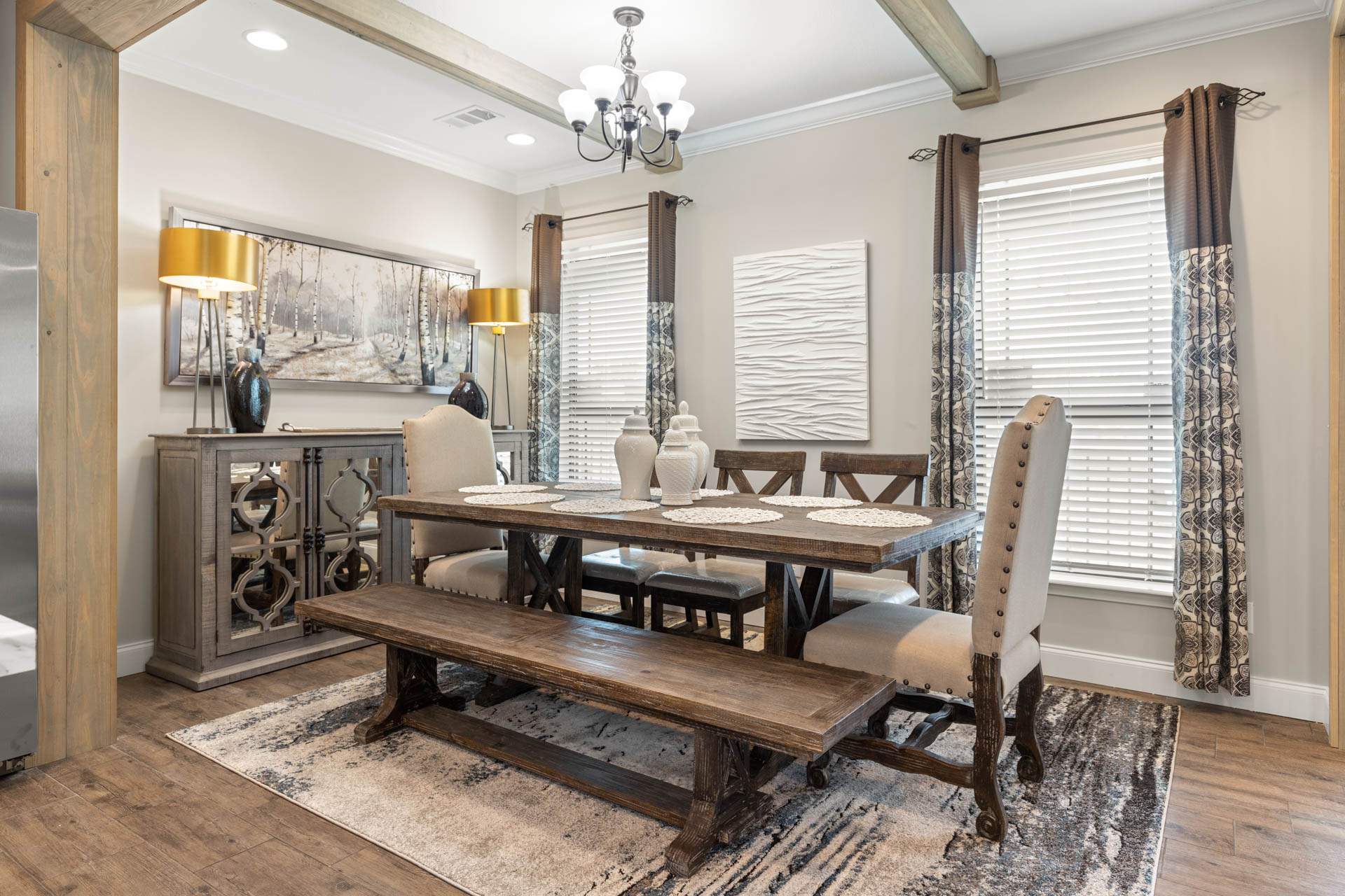 Elegant dining room with a rustic wooden table and bench, cushioned chairs, two vases, and a textured rug. Warm lighting from chandeliers and lamps create a cozy ambiance.
