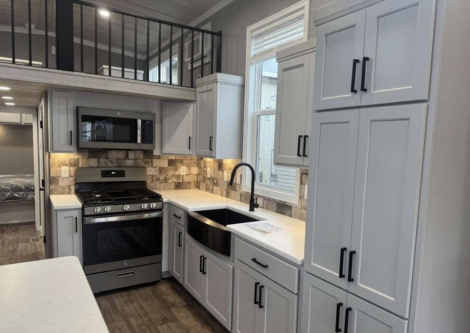 Modern kitchen with light gray cabinets, a black farmhouse sink, and stainless steel appliances. Warm lighting and wood floors add a cozy touch.
