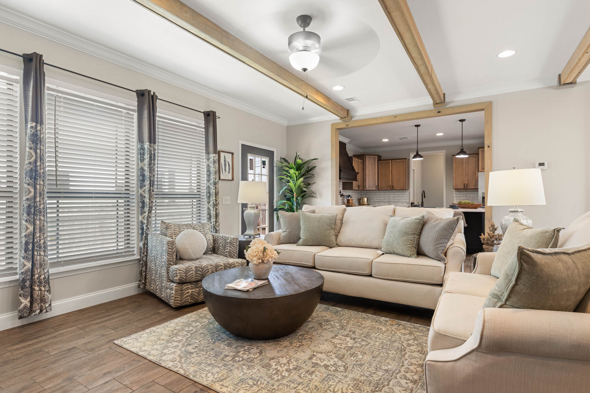 Stylish living room with beige sofas, patterned armchair, round coffee table on rug, and large windows. Wooden beams and plants create a cozy atmosphere.