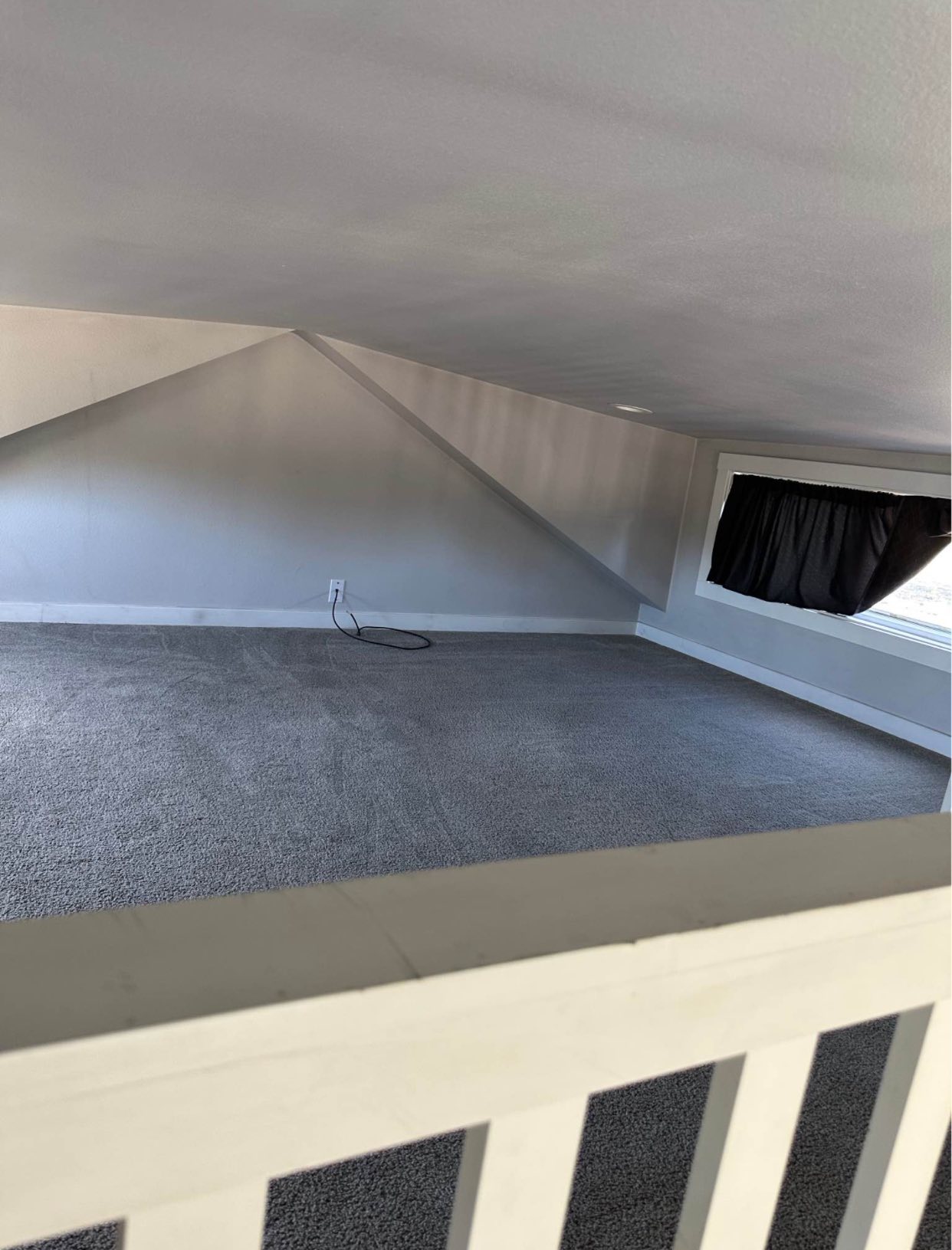 Small attic room with slanted ceiling, gray carpet, and a single window covered by a black curtain. The atmosphere feels cozy yet confined.