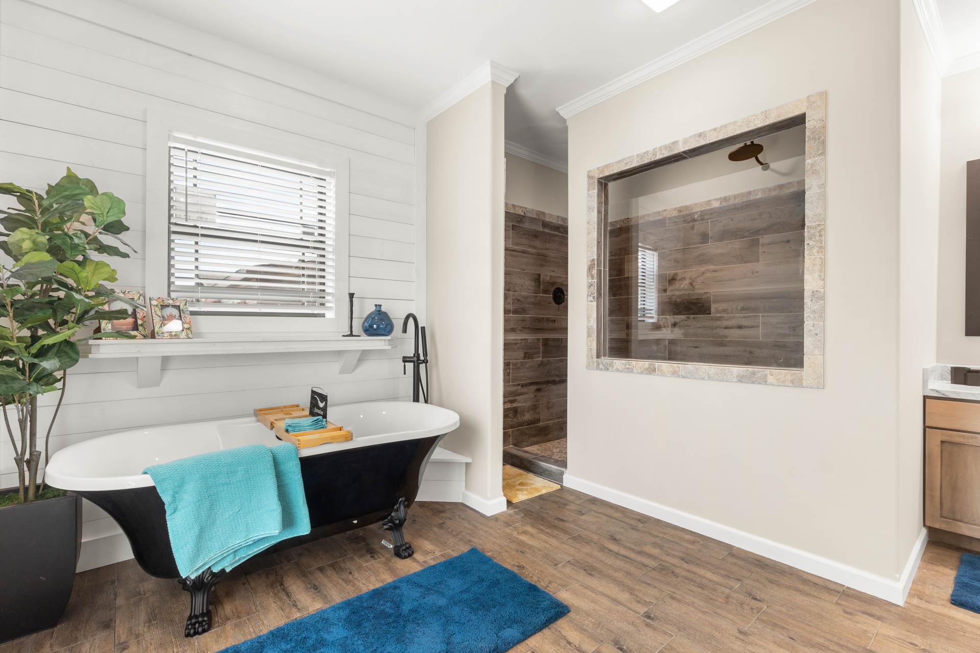 Bright bathroom with a freestanding black and white clawfoot tub, blue towel, wooden tray, and potted plant. Adjacent is a wooden-floored shower.