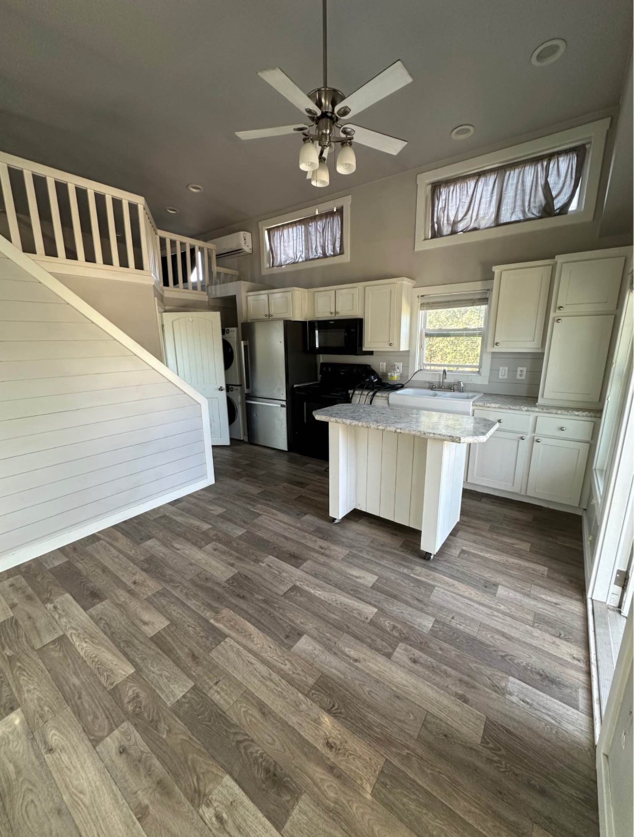 Cozy tiny house kitchen with light wood floors, white cabinets, and loft. Features include an island, black appliances, and a ceiling fan.