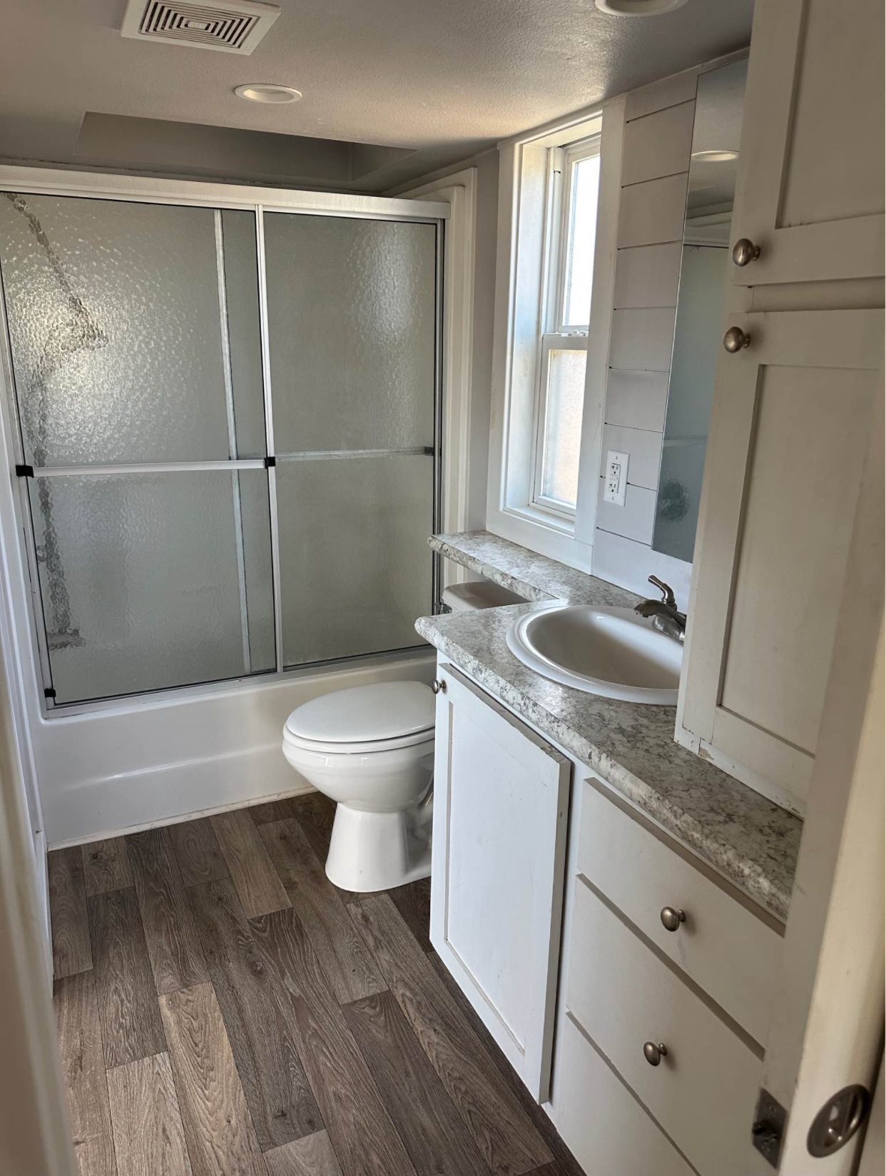 A small, modern bathroom with wood flooring, a shower with frosted glass doors, white toilet, and a granite countertop sink beside a window.