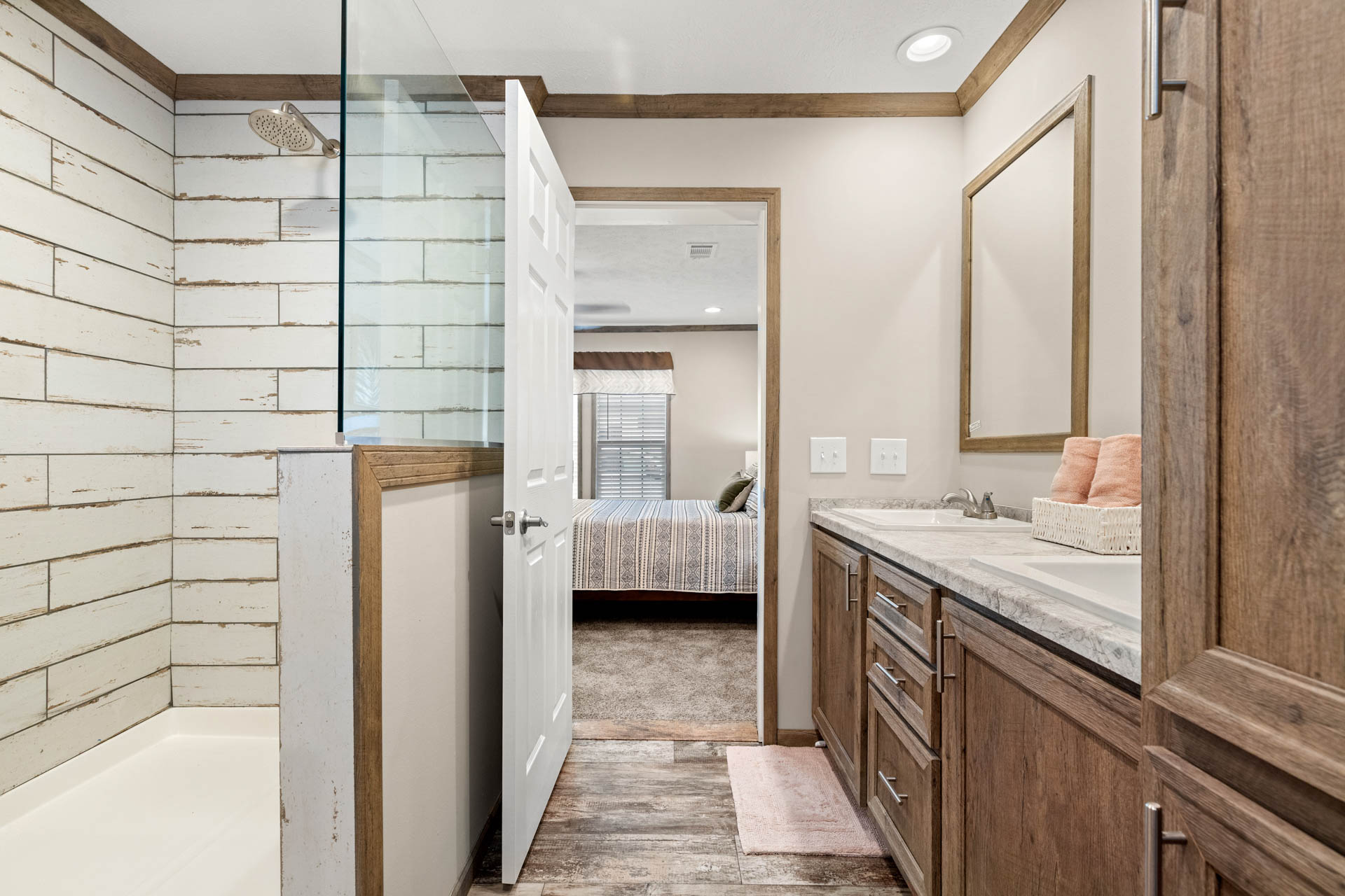 Modern bathroom with wood accents, featuring a glass shower on the left, a vanity with dual sinks on the right, and an open door leading to a bedroom.