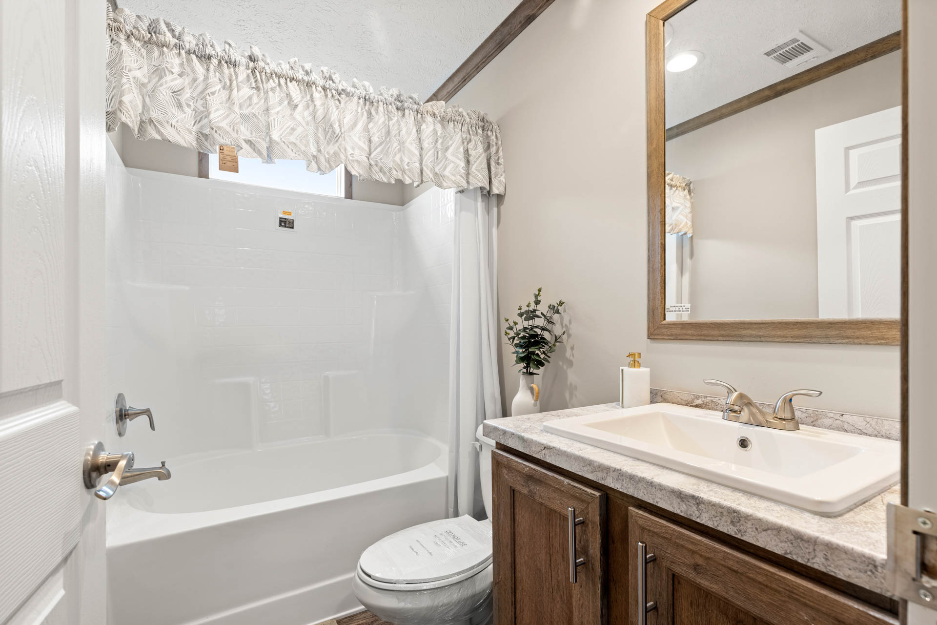 A bright, modern bathroom features a white bathtub with a patterned curtain, a stone countertop with a sink, wooden cabinets, and a mirror. The atmosphere is clean and inviting.
