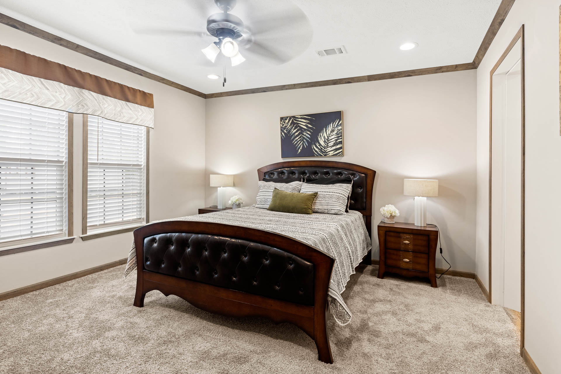 A cozy bedroom features a dark wooden bed with button-tufted leather accents, flanked by two bedside tables with lamps. A fan spins above, and artwork adorns the wall.