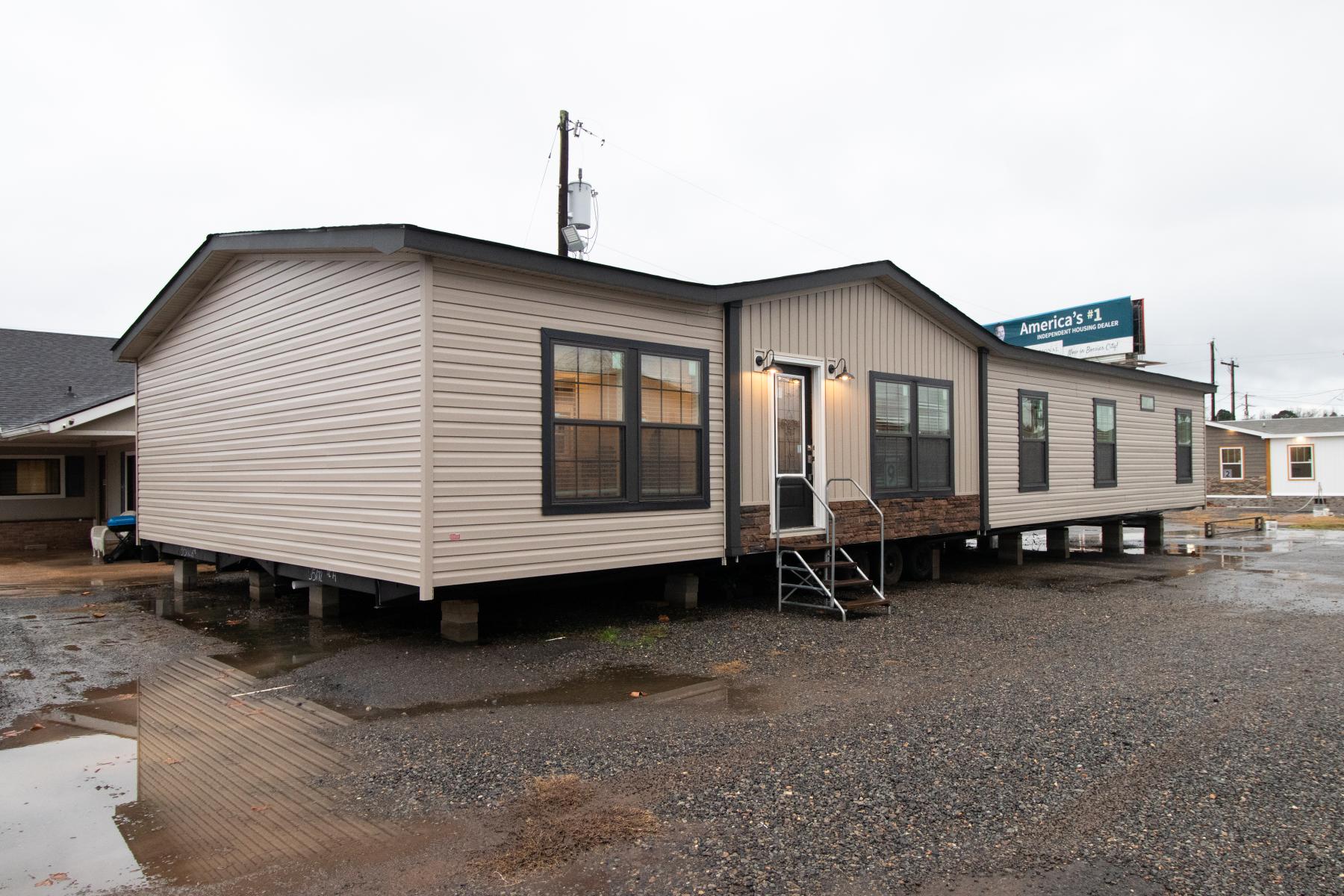A beige manufactured home raised on cinder blocks with a small metal staircase at the entrance, set against a cloudy sky, on a gravel lot with puddles.