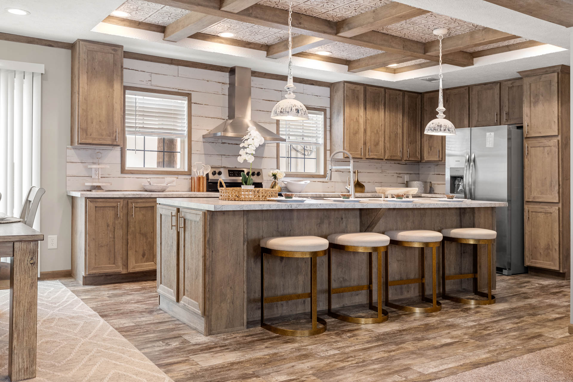 Rustic kitchen with wooden cabinets, central island, and four stools. Stainless steel appliances under pendant lights create a warm, inviting atmosphere.