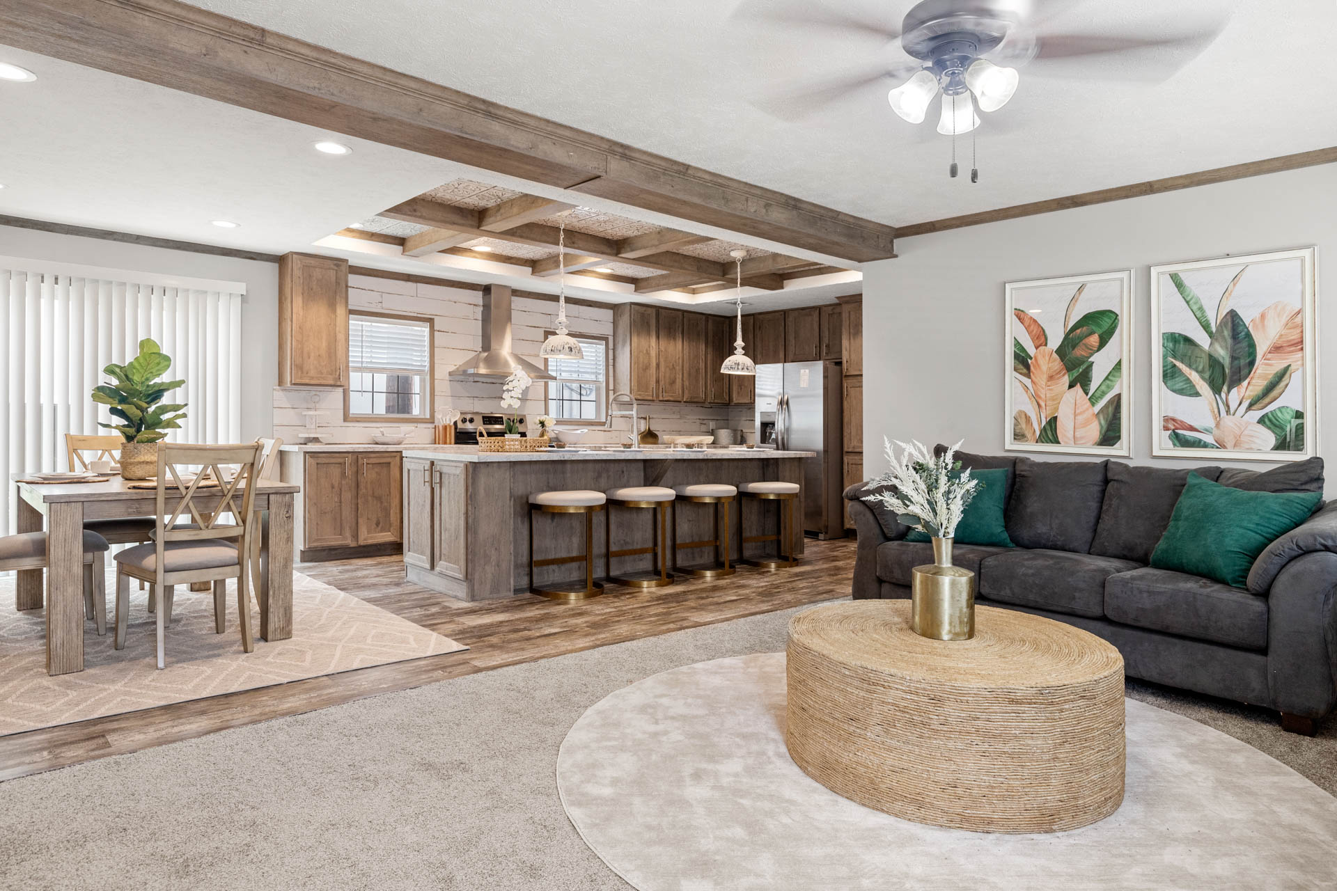Open-concept living area with a gray sofa, green cushions, and a woven coffee table. Wooden kitchen cabinets and a dining table create a cozy, modern feel.
