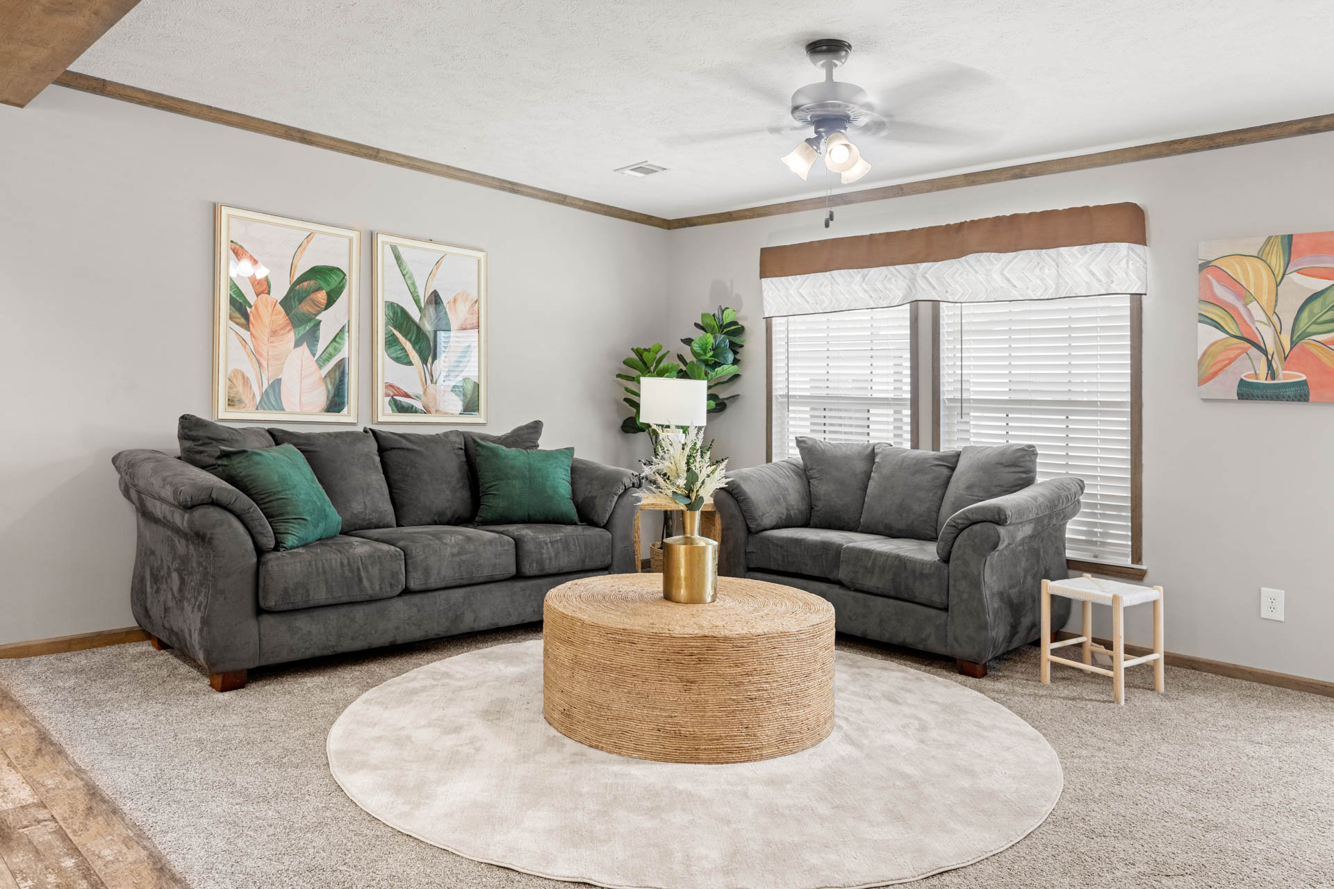 Cozy living room with two gray sofas against beige walls, tropical leaf artwork, a round wicker coffee table on a circular rug, and green cushions.
