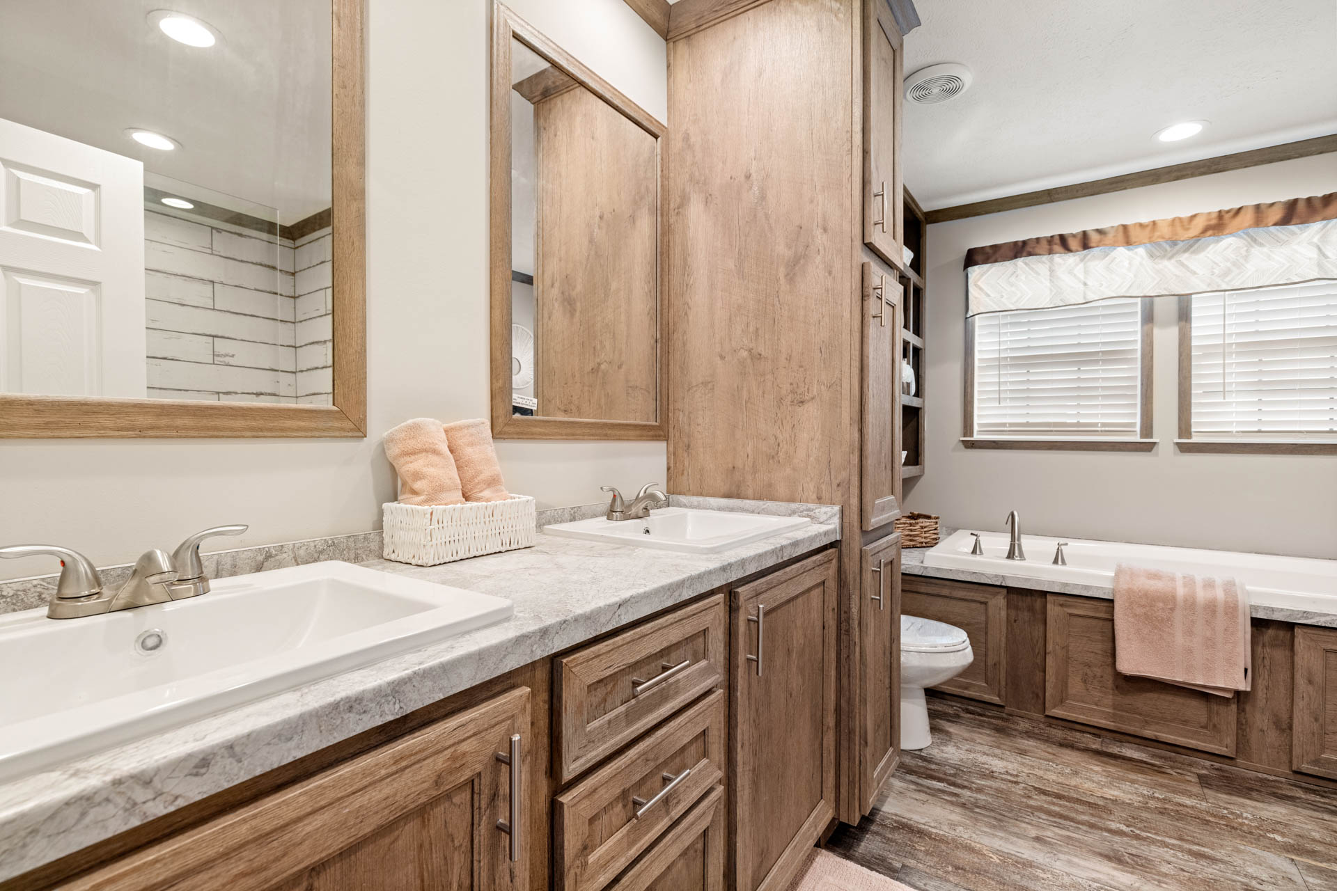 A cozy bathroom with wood cabinets, dual sinks, and a marble countertop. A bathtub with a peach towel is under two windows with white blinds.
