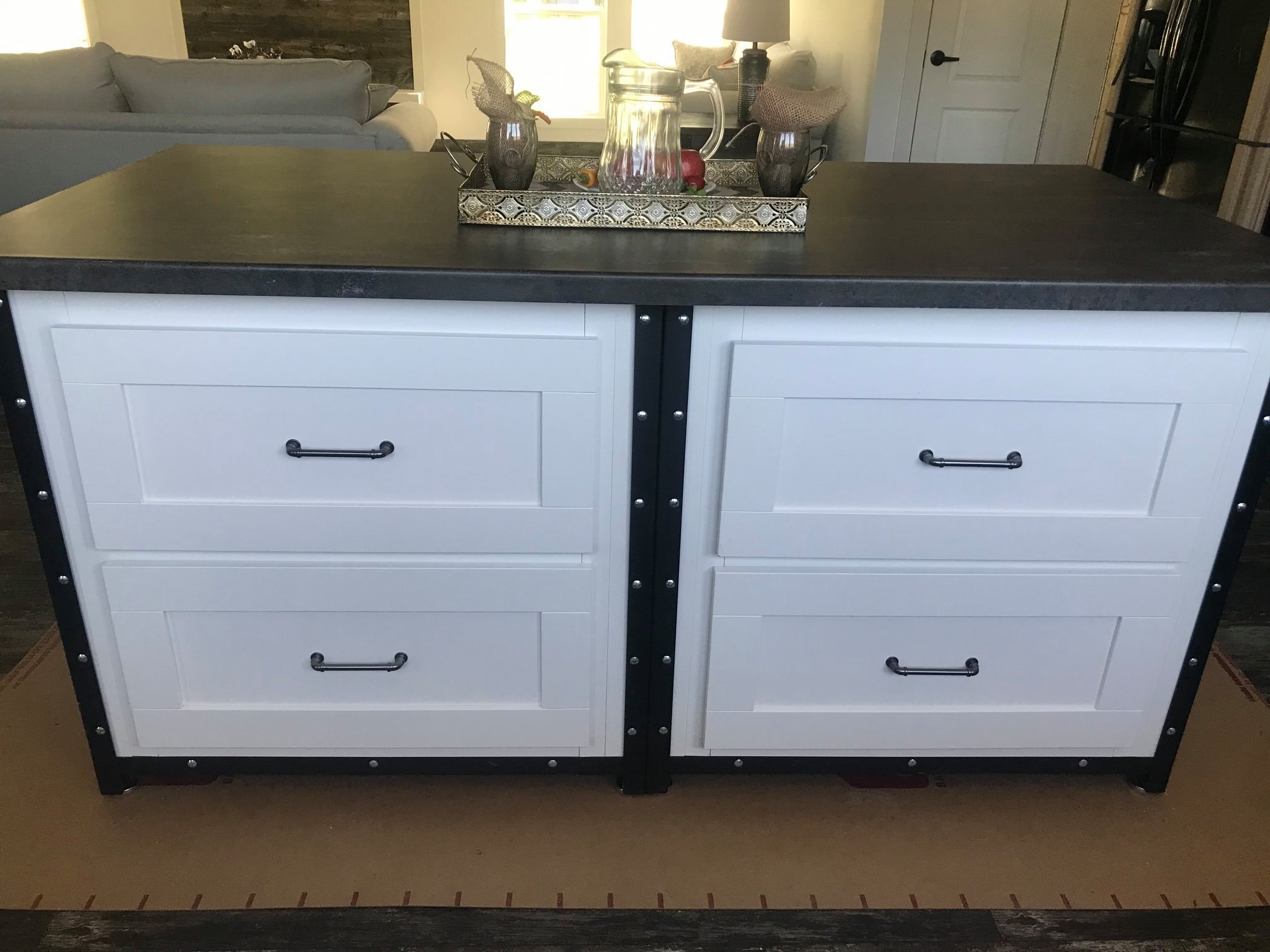 A white kitchen island with four drawers, black handles, and a dark countertop. A decorative tray on top holds glass jars and a fruit assortment. The background includes a living room with a gray couch.