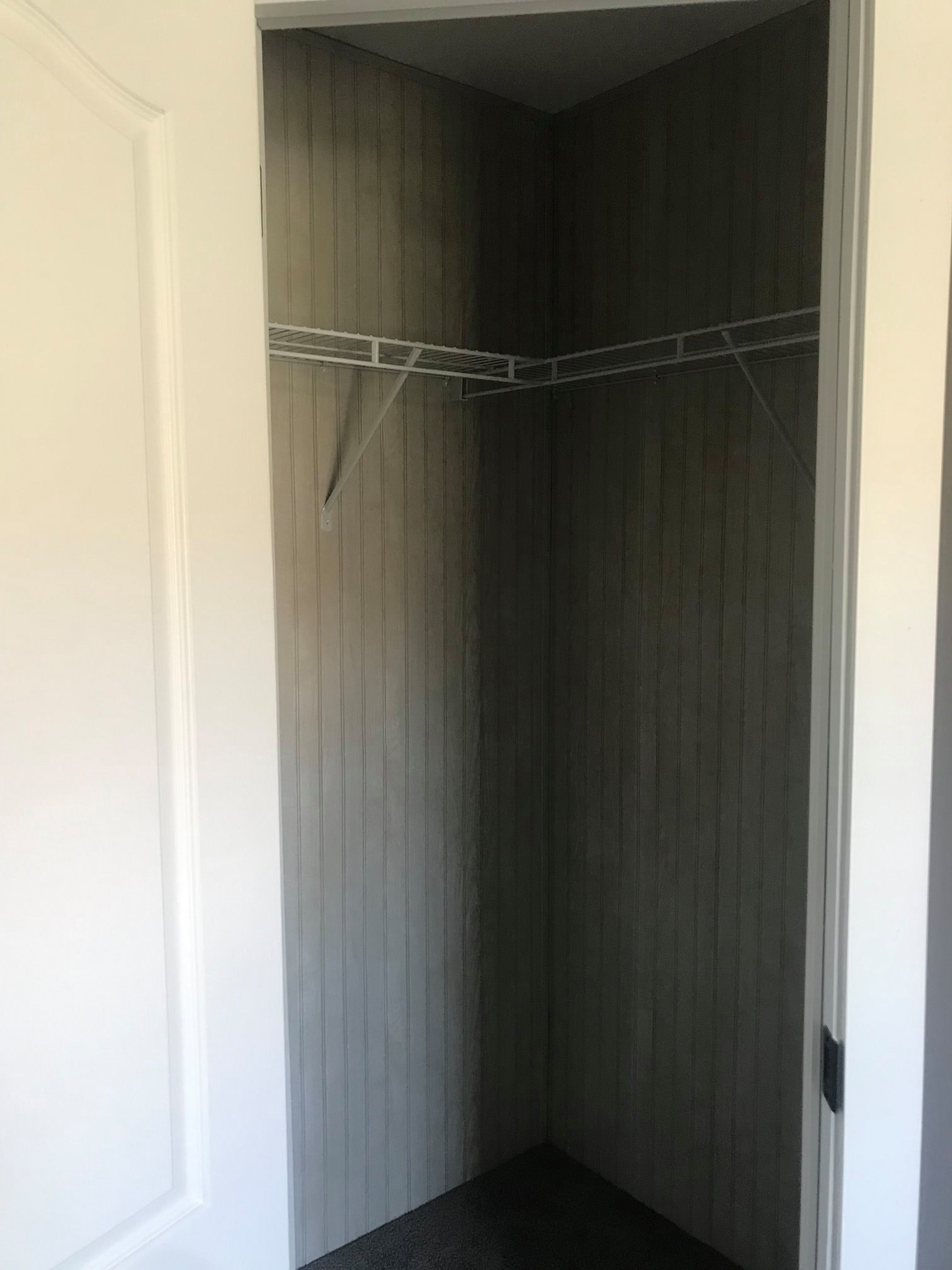 Compact, empty corner closet with open white door, featuring beige vertical wood paneling and a white metal wire shelf. Bright and minimalist.