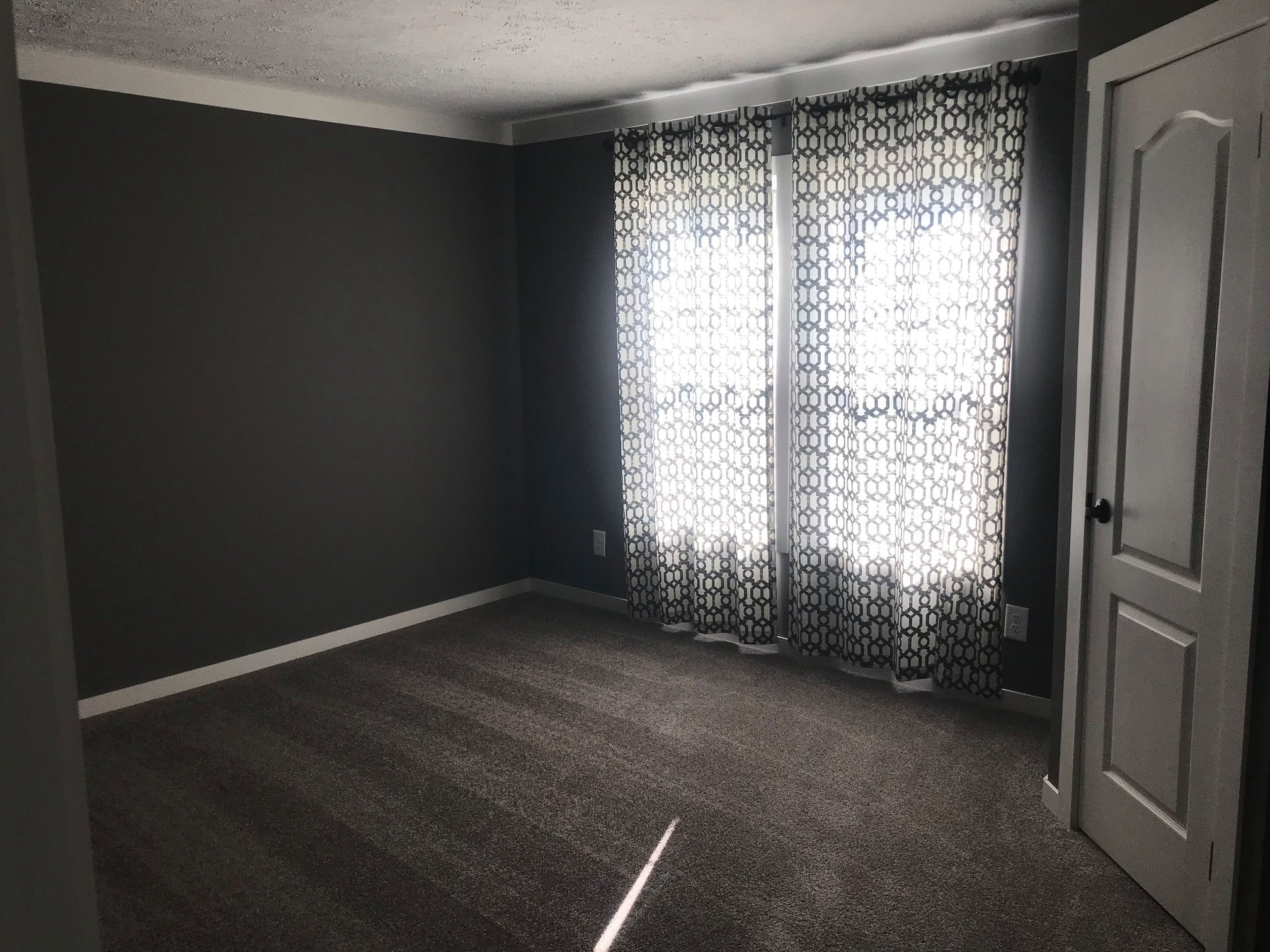Empty gray room with patterned curtains covering a window. Light filters through, creating a serene ambiance. White door on the right side.